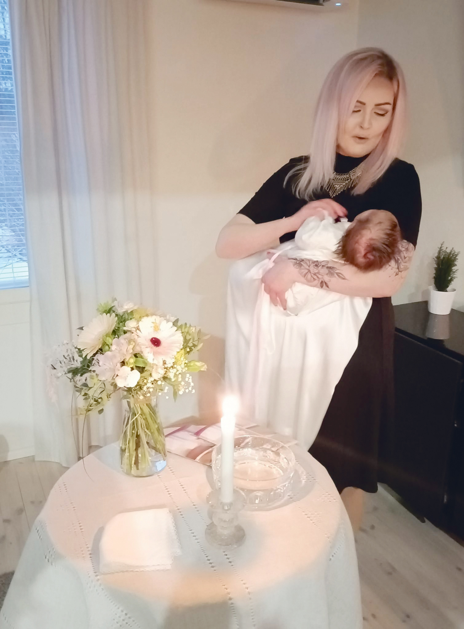 Naisella on vauva sylissä. Vauvalla on valkoinen kastemekko. Etualalla pöydällä on kastemalja, kukkakimppu ...