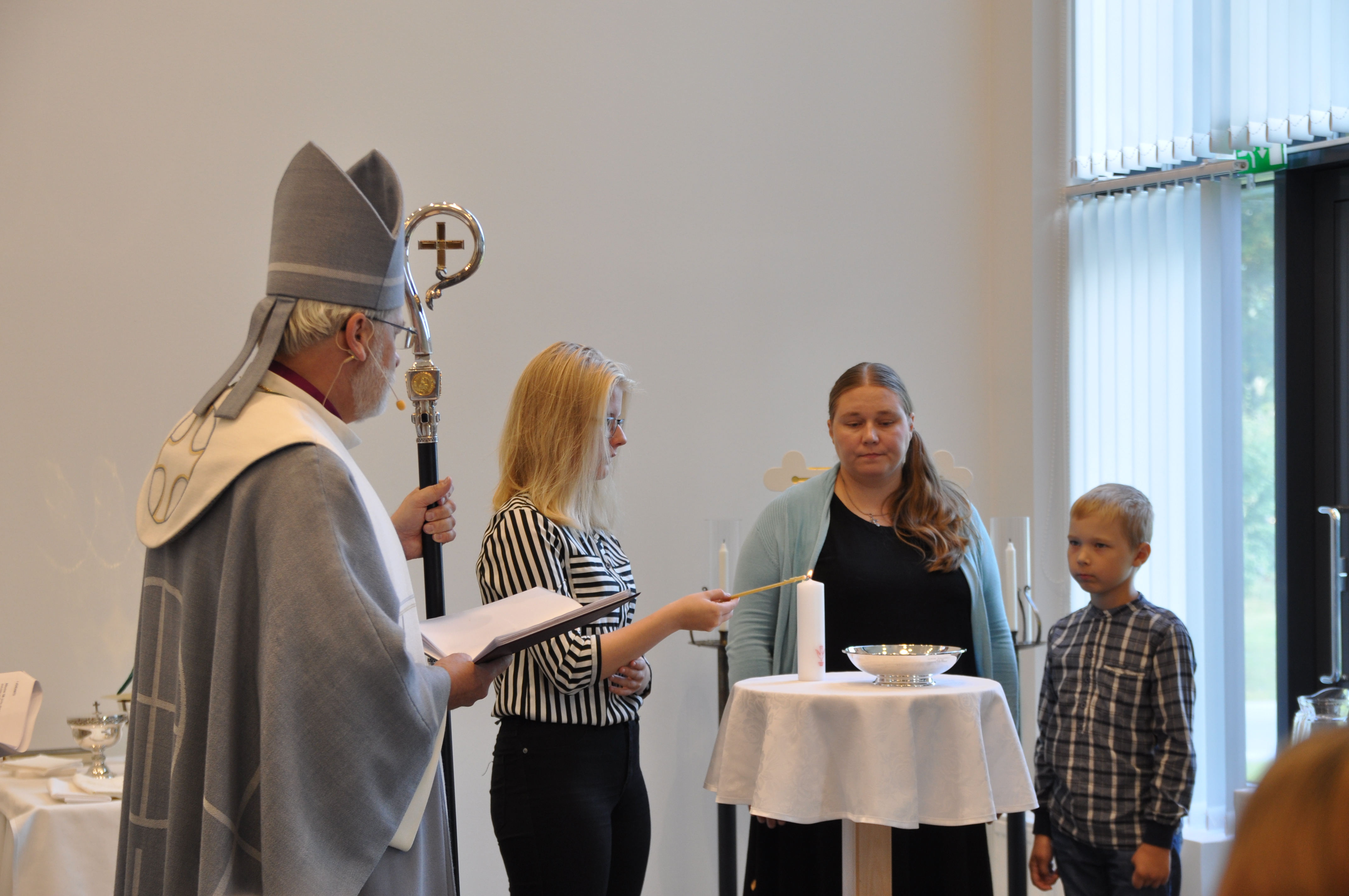 Kuvassa on neljä henkilöä, joista vasemmassa reunassa olevalla on piispan hiippa ja sauva. Keskellä on pöyt...