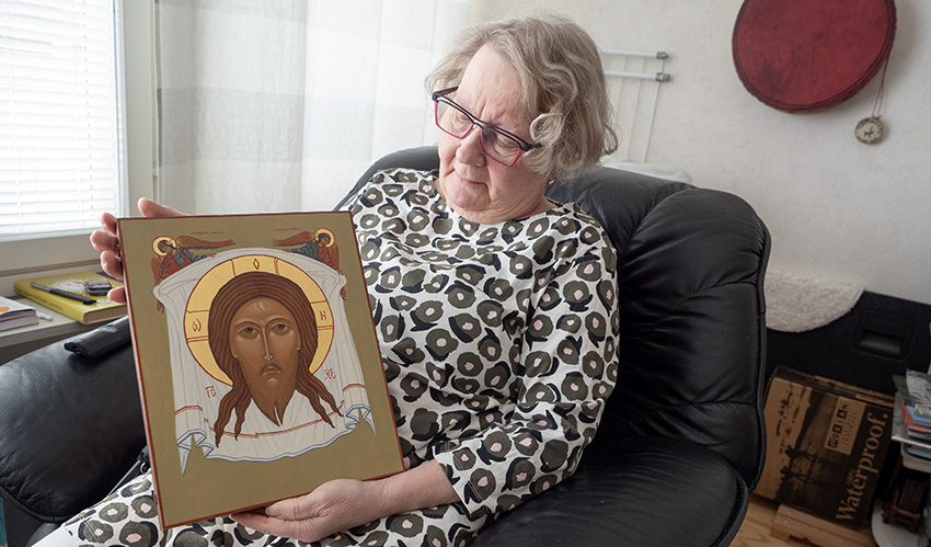 Vanhempi nainen istuu tuolissa ja pitää sylissään ikonia, jossa on Kristuksen kasvot kuvattuna liinaan.