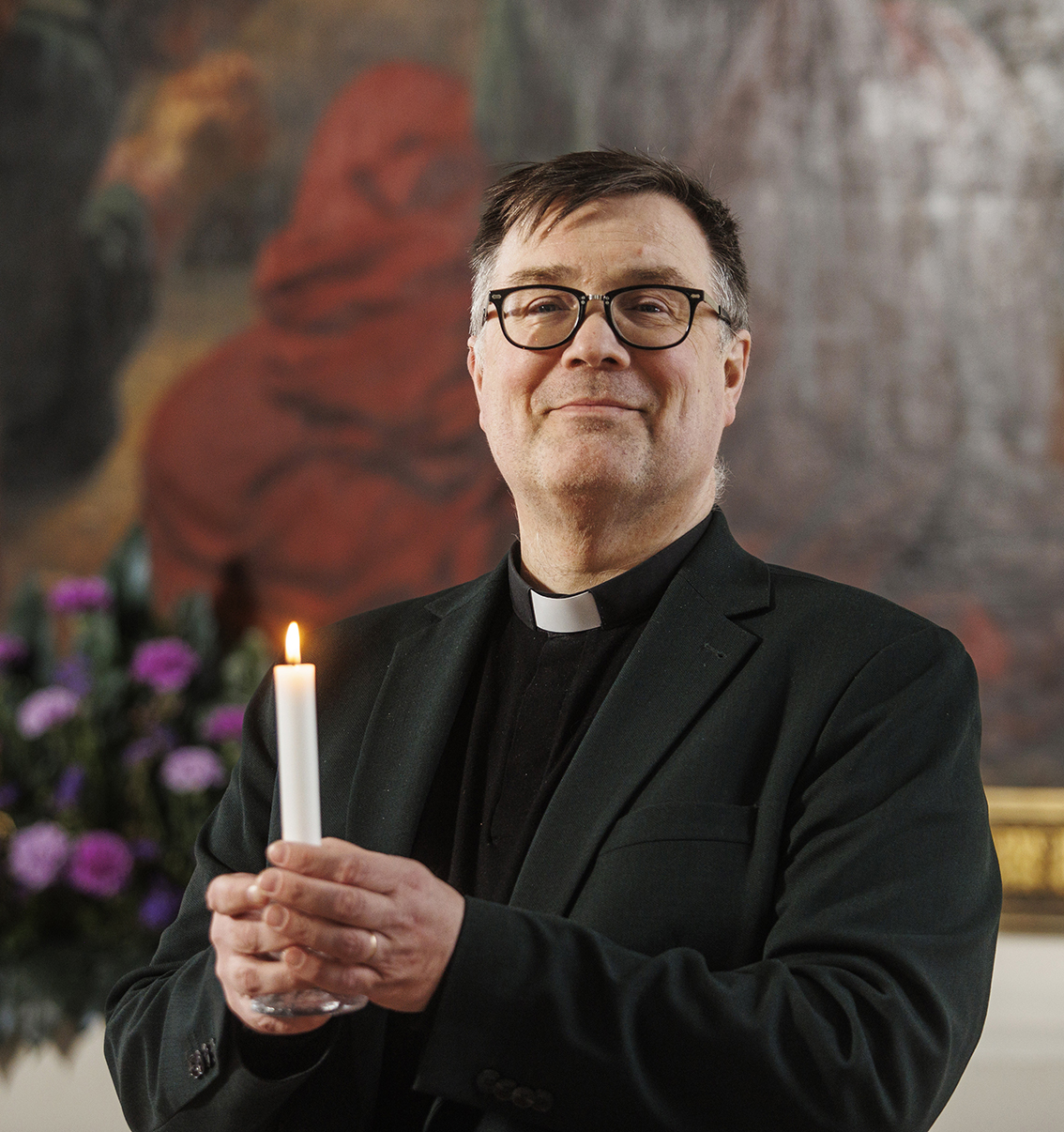 Pappi seisoo alttaritaulun edessä palava kynttilä kädessään.
