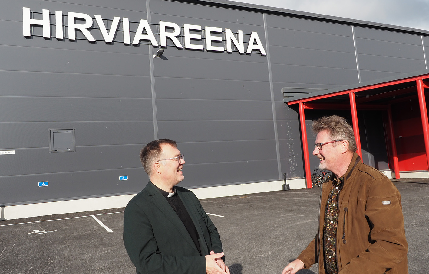 Taustalla iso halli, jonka seinässä lukee Hirviareena. Etualalla kaksi miestä, jotka katsovat toisiaan ja n...