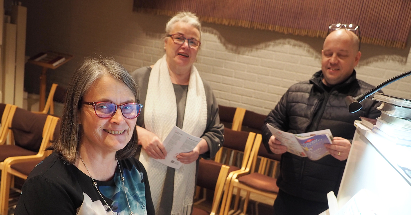 Kolme ihmistä pianon ääressä, kahdella lauluvihko kädessä.
