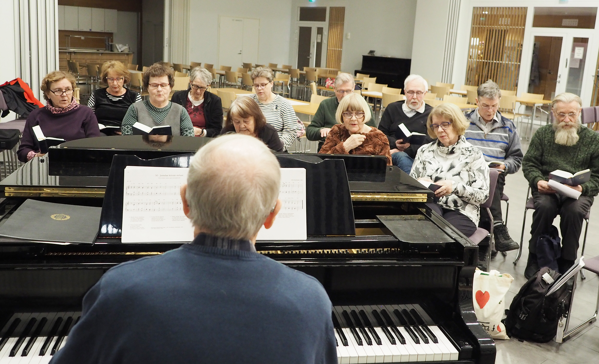 Mies soittaa pianoa, kuorolaiset istuvat pianon toisella puolella ja laulavat.