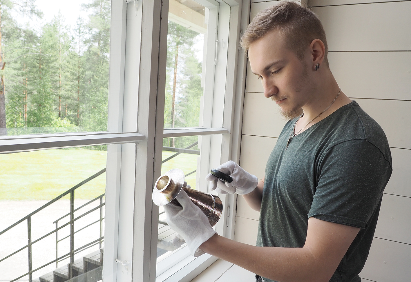 Mies pitää kädessään hopeista kynttilänjalkaa. Toisessa kädessä hänellä on suurennuslasi, jonka avulla mies...