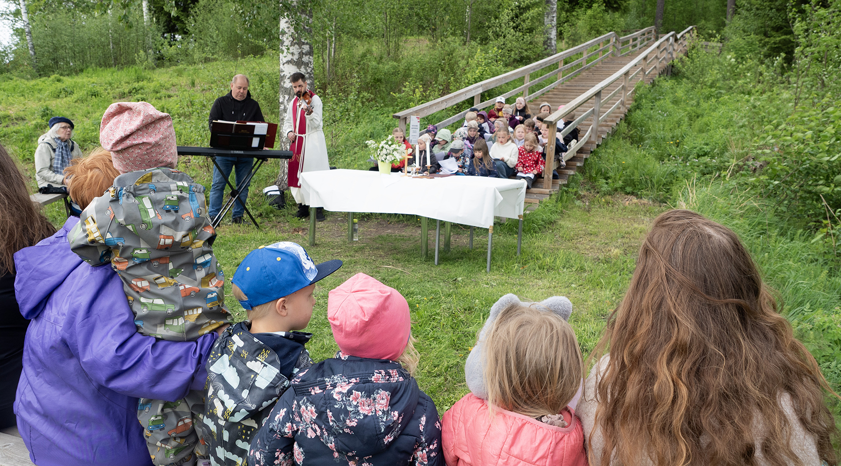 Etualalla äiti ja lapsia, taustalla kanttori sähköurkujen ääressä ja albaan pukeutunut pappi, joka soittaa ...