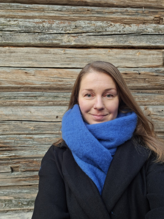 Nainen, jolla on pitkä, tumma tukka ja iso kaulahuivi kaulan ympärillä. Taustalla hirsiseinä.