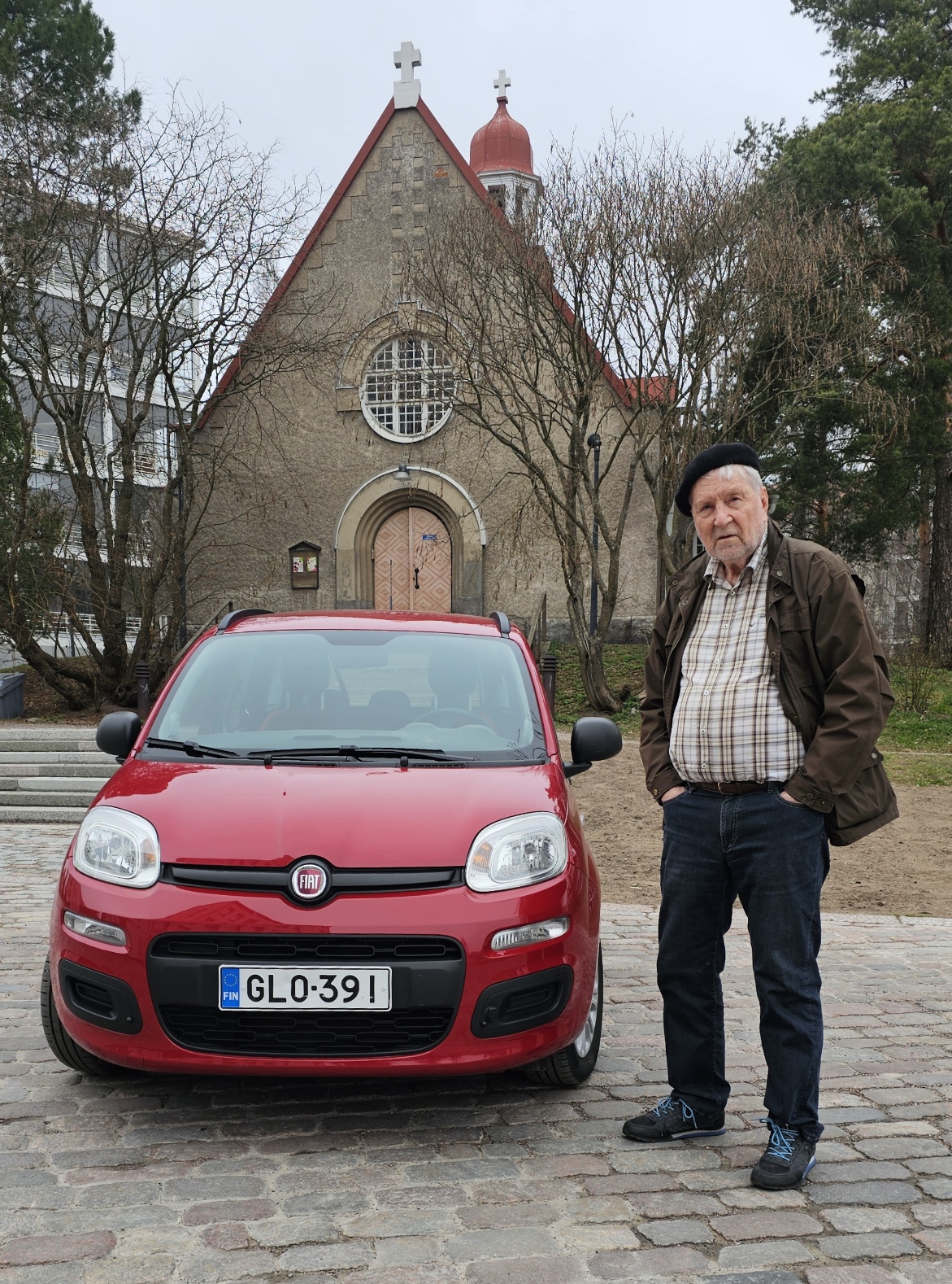 Mies seisoo punaisen pikkuauton vieressä, taustalla kivikirkko.
