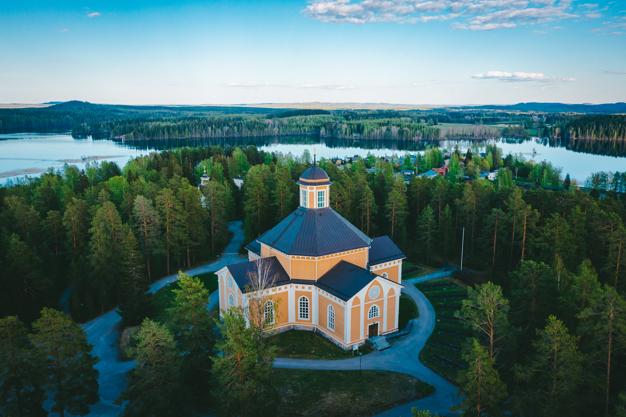 Keltainen puuristikirkko metsän keskellä, taustalla näkyy järvi.